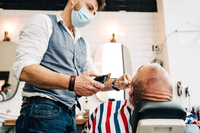 D'en bas maître coiffeur sur le visage masque médical barbe de coupe de l'homme masculin avec machine électrique dans le salon de coiffure pendant la pandémie covid — Photo de stock