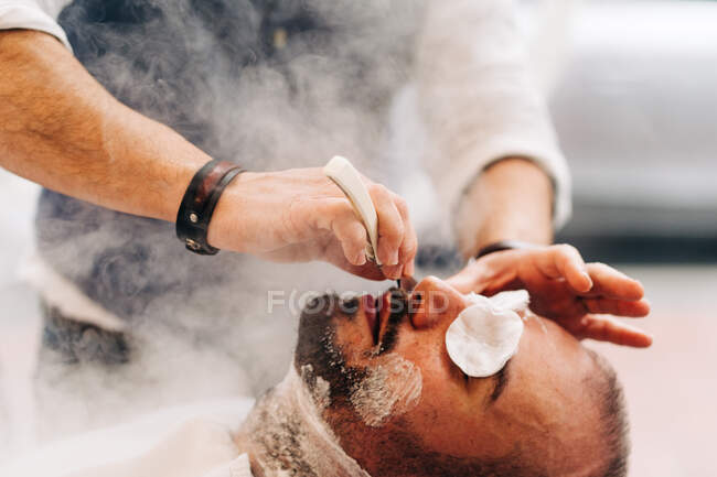 Crop anonymous beauty master shaving beard of client with straight razor during steam vapor treatment in hairdressing salon - foto de stock