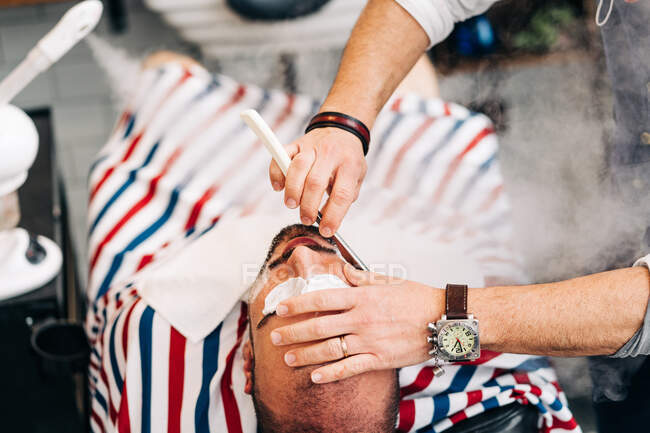 From above of crop anonymous beauty master in wristwatch shaving beard of client with straight razor during steam vapor treatment in hairdressing salon — Stock Photo