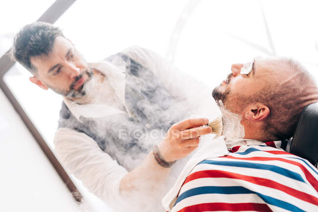 From below male beauty master shaving beard of client with straight razor during steam vapor treatment in hairdressing salon — Fotografia de Stock