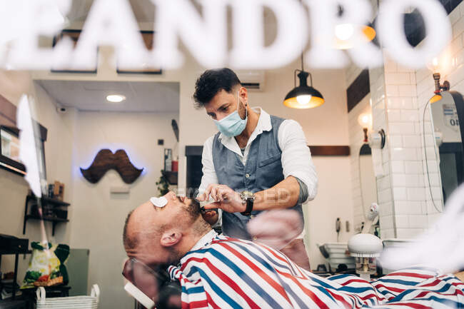 Through glass wall view of anonymous masculine stylist shaving beard of man with cotton pads on eyes using straight razor in beauty salon - foto de stock