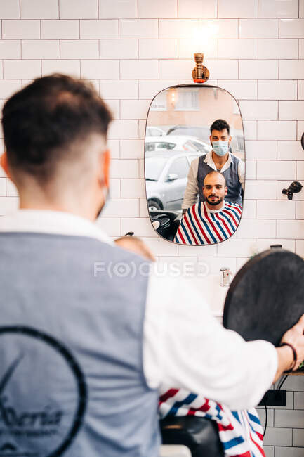 Вид ззаду анонімного чоловічого перукаря в масці біля бородатого клієнта, що відображає в дзеркалі в перукарні — стокове фото