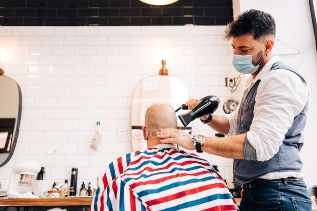 Anonymous male stylist in mask using hairdryer while touching bald head of man in striped cloth in beauty salon - foto de stock