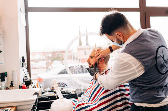 Back view of anonymous masculine stylist barber on face mask shaving beard of man using straight razor in beauty salon during coronavirus pandemic - foto de stock
