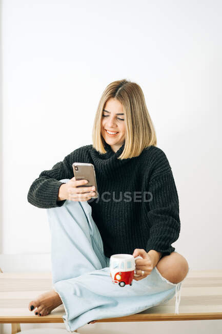 Junge zufriedene Barfußweibchen mit einer Tasse Getränk surfen auf dem Handy im Internet, während sie sich im Haus auf weißem Hintergrund ausruhen — Stockfoto