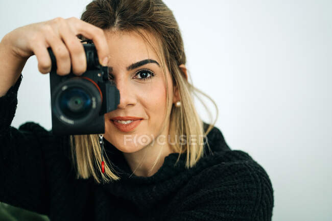 Кадрирование улыбается женщина в черном свитере глядя на камеру во время фотографирования на профессиональную цифровую камеру в доме на белом фоне — стоковое фото