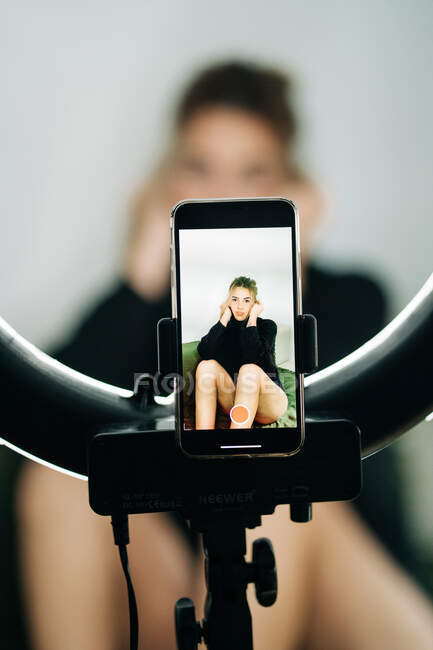 Junge barfüßige Frau berührt Haare und Wangen auf Couch, während sie zu Hause durch Lichtring mit Handy in die Kamera schaut — Stockfoto