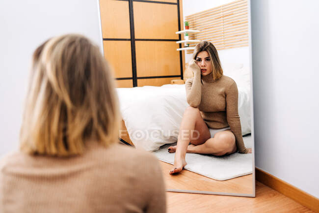 Visão traseira da encantadora jovem pensiva tocando bochecha contra espelho enquanto sentado em parquet em casa — Fotografia de Stock