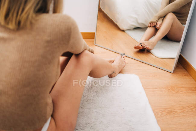 Back view of young pensive female touching leg against mirror while sitting on parquet in house — Stock Photo