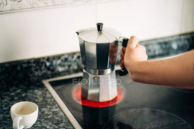 Crop person holding metal stove top coffee maker with plastic handle on modern hot hob in house kitchen in daylight — Stock Photo