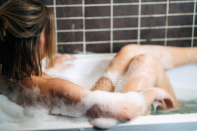 Vista lateral da jovem fêmea sedutora deitada na banheira com espuma na casa — Fotografia de Stock