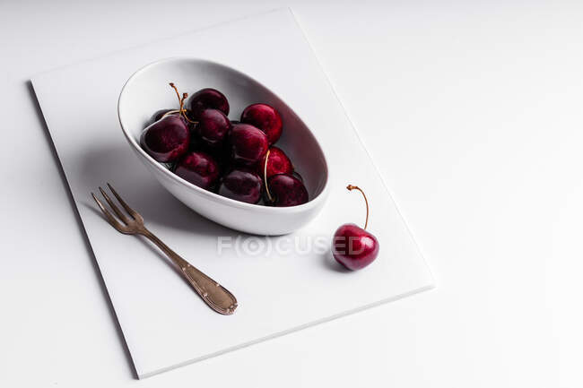 High angle of healthy ripe tasty cherries in ceramic bowl placed with fork on white board — Stock Photo