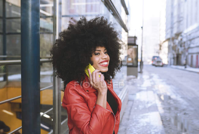 Vista lateral de la mujer afroamericana feliz con el pelo rizado sonriendo ampliamente mientras habla en el teléfono inteligente en la calle de la ciudad - foto de stock
