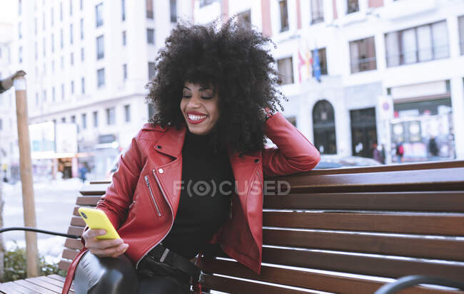 Stylish astonished African American female reading news on cellphone sitting on bench in city — Stock Photo