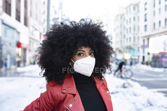 Serious African American female with curly hair in protective mask standing on street covered with snow — Stock Photo