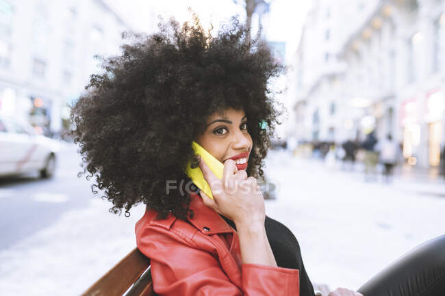 Visão lateral da mulher afro-americana feliz com cabelo encaracolado sorrindo amplamente enquanto fala no smartphone na rua da cidade — Fotografia de Stock