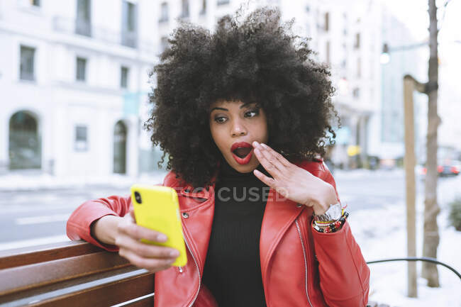 Stylish astonished African American female reading news on cellphone with mouth opened sitting on bench in city — Stock Photo