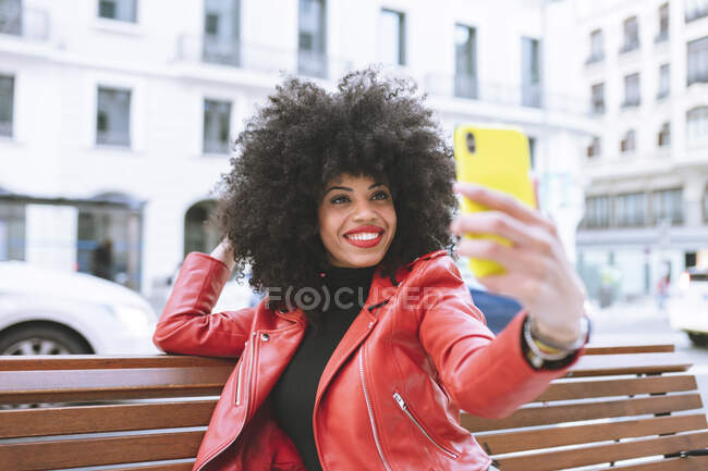 Foco seletivo da mulher afro-americana feliz com penteado afro sentado no banco e tomando auto-retrato no celular — Fotografia de Stock