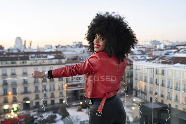 Back view of African American female with curly hair in stylish outfit standing on balcony and looking at city buildings — Stock Photo