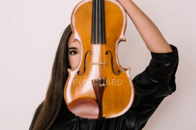 Künstlerin bedeckt Gesicht mit Geige, während sie vor weißem Hintergrund steht und in die Kamera schaut — Stockfoto