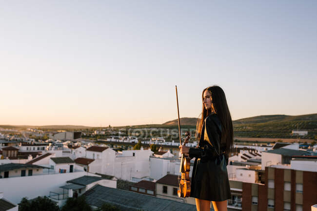 Rückseite junge schöne Musikerin in stylischem Minikleid mit akustischer Geige steht auf dem Dach eines Wohnviertels und schaut an sonnigen Abenden weg — Stockfoto