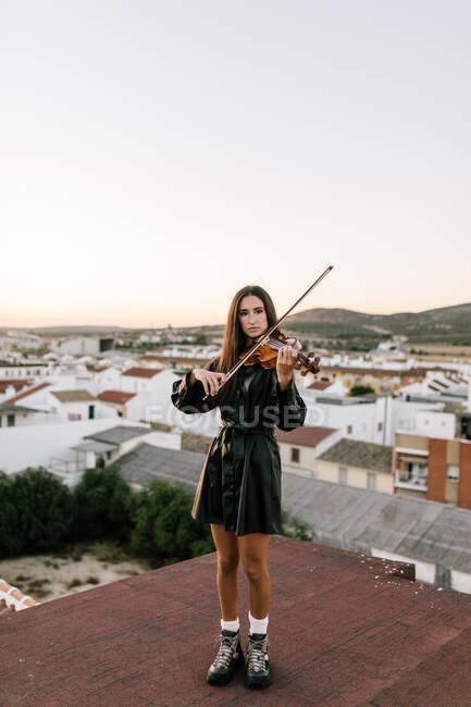 Молодая красивая женщина-музыкант в стильном мини-платье держит акустическую скрипку и стоит на крыше в жилом пригороде и смотрит в солнечный вечер — стоковое фото
