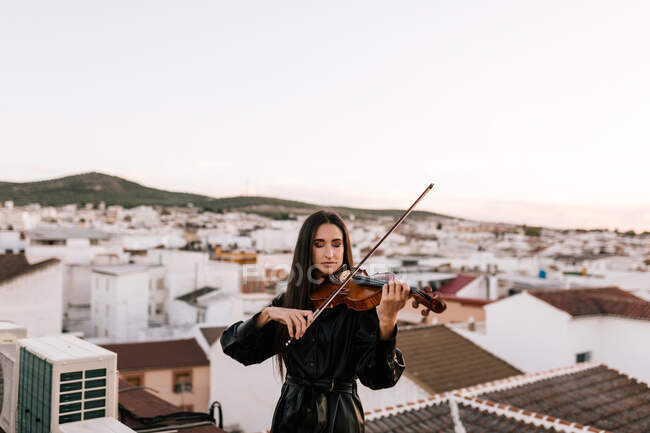 Молодая красивая женщина-музыкант в стильном мини-платье держит акустическую скрипку и стоит на крыше в жилом пригороде и закрывает глаза в солнечный вечер — стоковое фото