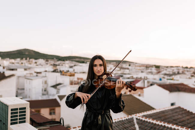 Junge schöne Musikerin in stylischem Minikleid mit akustischer Geige steht auf dem Dach eines Wohnviertels und blickt an einem sonnigen Abend in die Kamera — Stockfoto