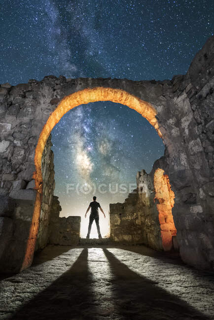 Vista posterior silueta de viajero anónimo de pie en la antigua iglesia en ruinas y admirando la noche estrellada en España - foto de stock