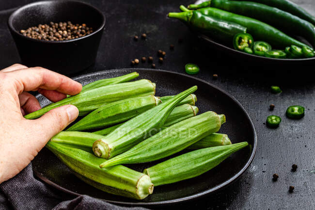 Cultures personne méconnaissable tenant okra mûr sur la table avec des légumes frais sur la poêle — Photo de stock