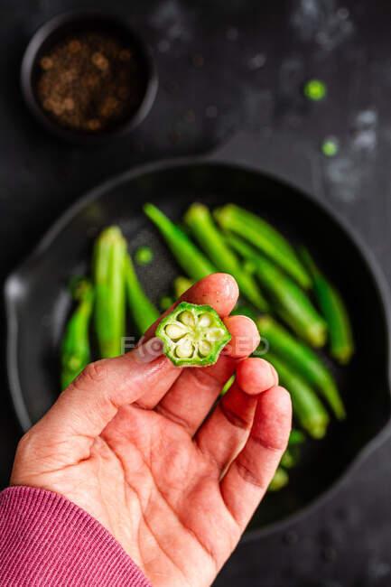 Из выше урожая неузнаваемый человек держит спелую окру над столом со свежими овощами на сковороде — стоковое фото