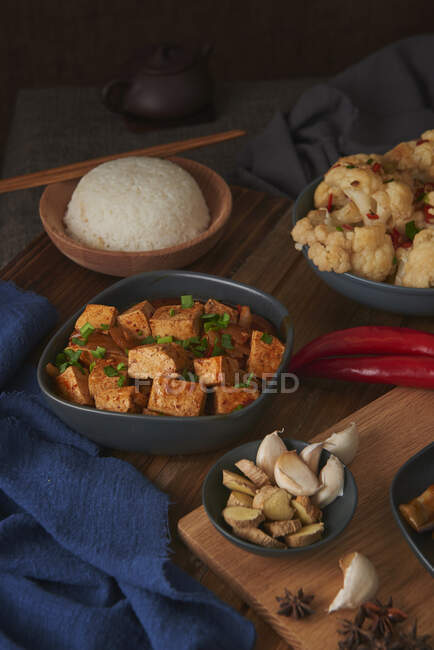 Mala tofu, plato vegano chino, acompañado de un tazón de arroz, coliflor, salsa de soja y una tetera japonesa encima de una mesa de madera decorada con telas - foto de stock
