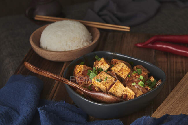 Закройте мала тофу, китайское веганское блюдо, в сопровождении миски риса и японского чайника на деревянном столе, украшенном тканями — стоковое фото