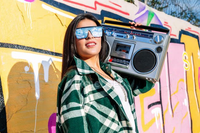 Jeune femme à la mode dans des lunettes de soleil reflétant ciel nuageux écouter des chansons de vieux magnétophone contre mur de graffiti à la lumière du soleil — Photo de stock