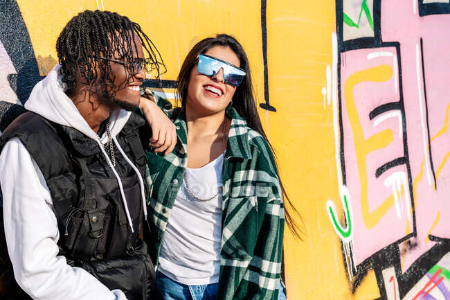 Jeune homme hipster noir joyeux près du contenu partenaire féminin dans les lunettes de soleil modernes passer du temps ensemble dans la rue à la lumière du soleil — Photo de stock