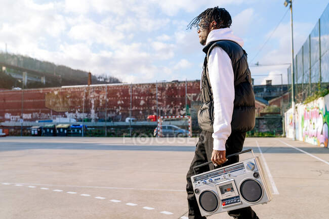 Side view of young African American male in trendy apparel walking on city street with old tape recorder in sunlight — Stock Photo