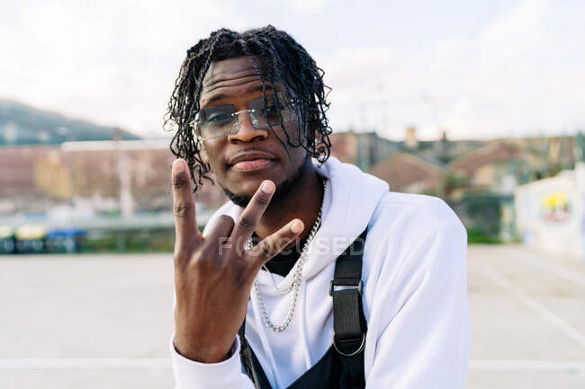 Homem afro-americano na moda em óculos de sol com tranças afro demonstrando gesto rap enquanto olha para a câmera na cidade — Fotografia de Stock