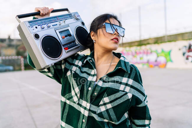 Young trendy female in sunglasses reflecting cloudy sky listening to song from old tape recorder against graffiti wall in sunlight — Stock Photo