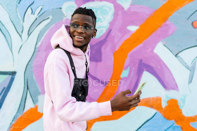 Side view of young content ethnic hipster male in trendy apparel and sunglasses with cellphone looking away in town — Stock Photo