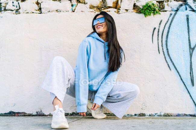 Young cheerful female in trendy apparel and modern sunglasses squatting on walkway in town in daylight — Stock Photo