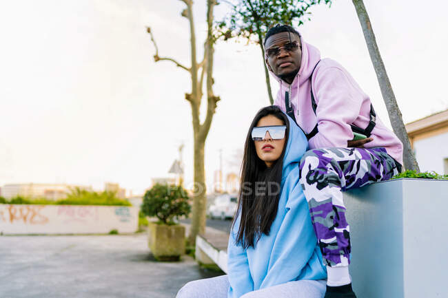 Молодой черный мужчина в модной одежде сидит рядом с женщиной-партнером в современных солнцезащитных очках и смотрит вдаль в город — стоковое фото