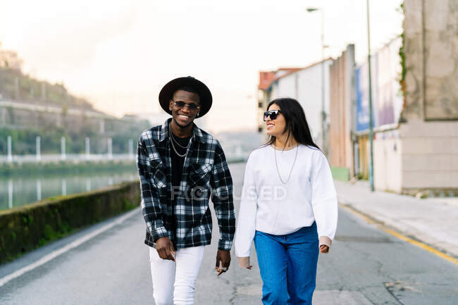 Young content multiracial partners in trendy clothes and sunglasses talking while looking at each other and strolling on city roadway - foto de stock