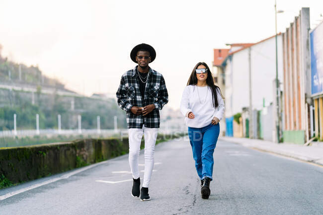 Молодой контент мультирасовые партнеры в модной одежде и солнцезащитных очках говорить при взгляде на камеру на городской проезжей части — стоковое фото