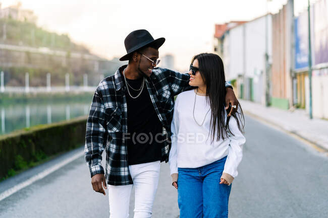 Young content multiracial partners in trendy clothes and sunglasses talking while looking at each other and strolling on city roadway - foto de stock