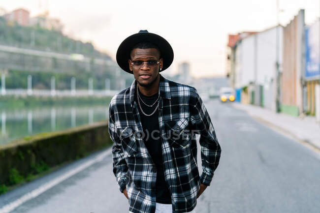 Young African American male in trendy wear and chain looking at camera on urban asphalt roadway - foto de stock