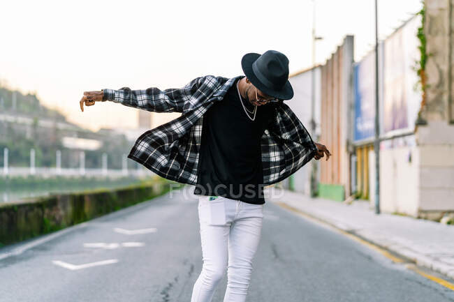 Young African American male in trendy wear and chain looking down on urban asphalt roadway - foto de stock