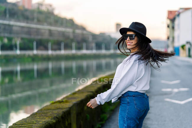 Side view of young happy female in trendy clothes and sunglasses dancing on road against water channel in city — Stock Photo