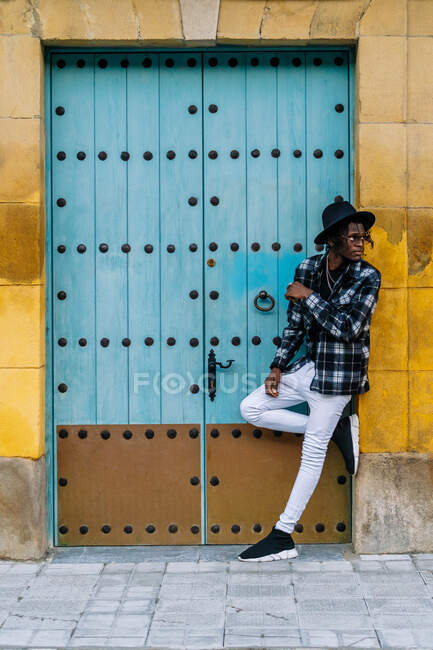 Full body of African American male in trendy outfit and sunglasses standing with crossed legs near doorway — Foto stock