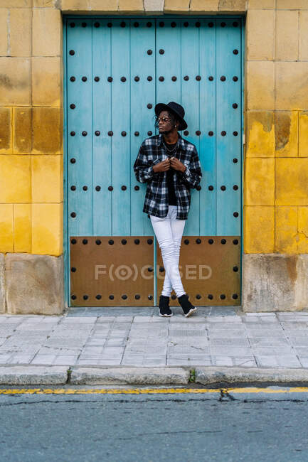 Повний корпус афроамериканського чоловіка в модних костюмах і сонцезахисних окулярах стоячи зі схрещеними ногами біля дверей. — стокове фото