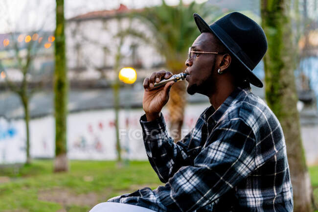Serious African American male wearing stylish checkered shirt sunglasses and hat smoking cigarette - foto de stock